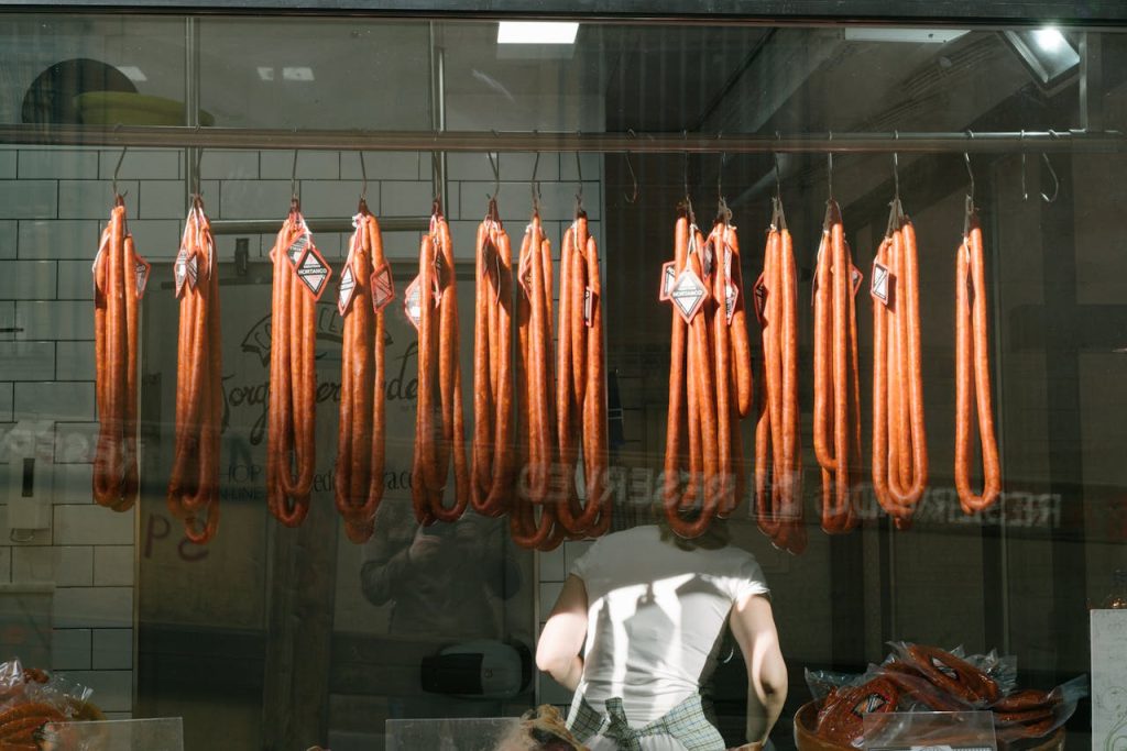 Through glass back view of unrecognizable worker in white T shirt at meat preserving factory with long sausages hanging from metal rail in tiled room